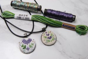 Two cross stitch pendants next to a skein of Cosmo Seasons and some spools of Nishikiito thread.