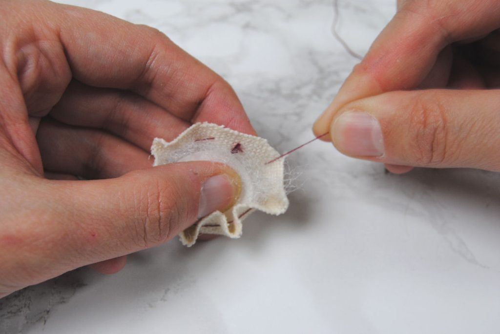 Demonstration of pulling a running stitch tight to cinch fabric around a cardstock circle.