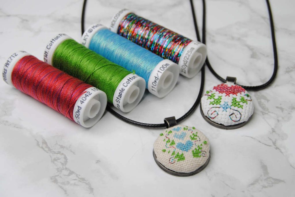Two Cross stitched pendants next to the spools of Sulky thread used to stitch them.