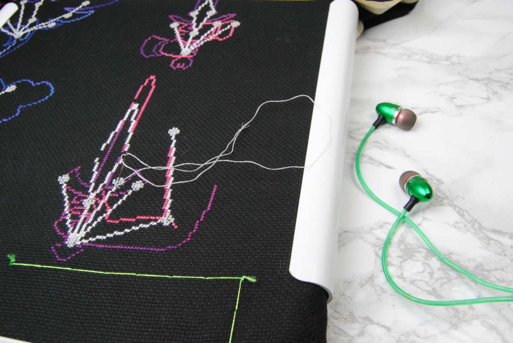 Close up of a cross stitch of the Skyrim skill constellations next to some earbuds.