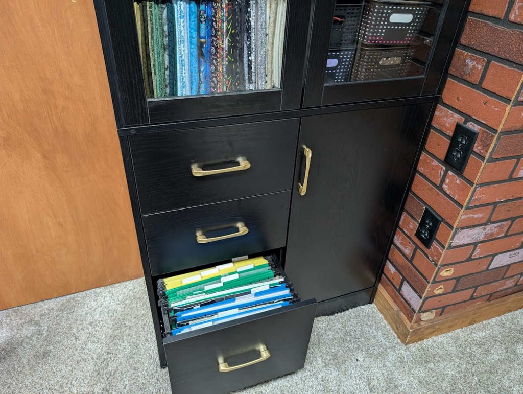 A tall cabinet with a file cabinet drawer being used to store cross stitch patterns, wips, and materials.