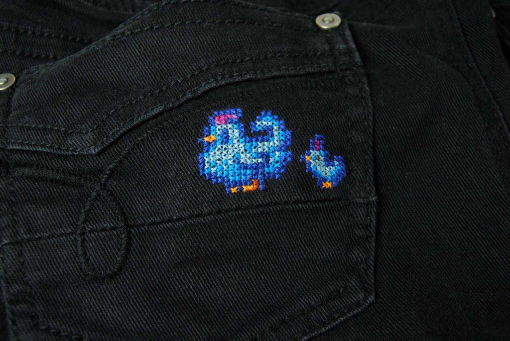 Finished Stardew Valley blue chicken cross stitch on the pocket of some black jean shorts.