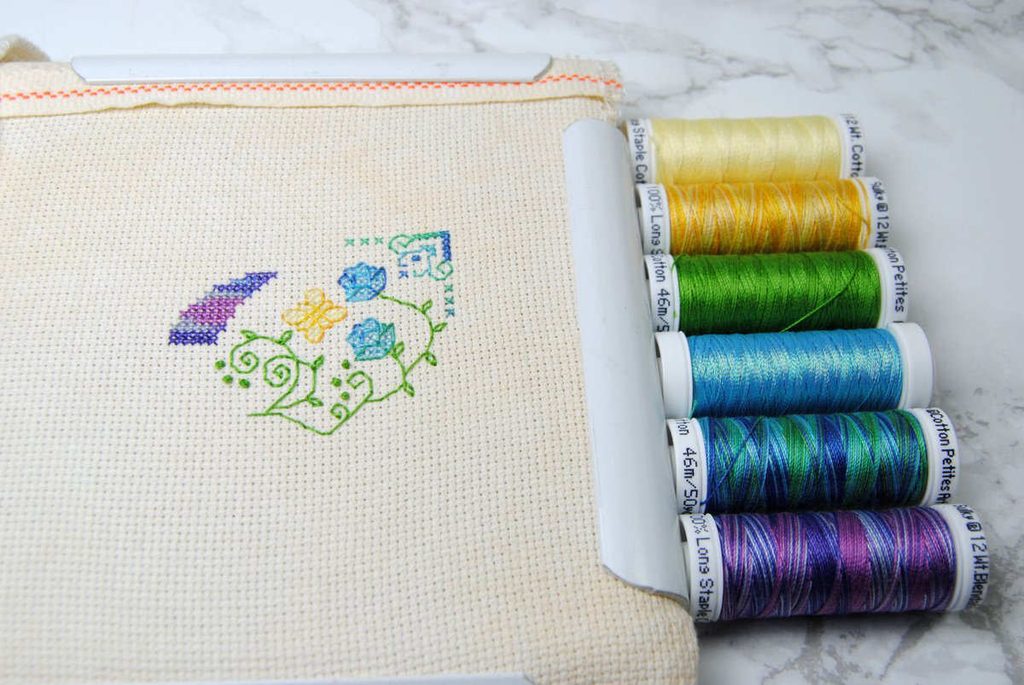 A cross stitch in progress next to 6 spools of Sulky thread.