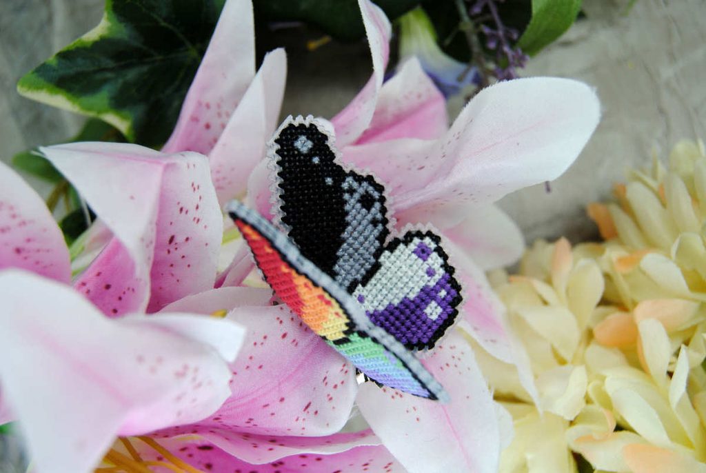 A 3d poseable cross stitch butterfly with pride flag wings. A pastel rainbow outside, and an asexual flag inside.