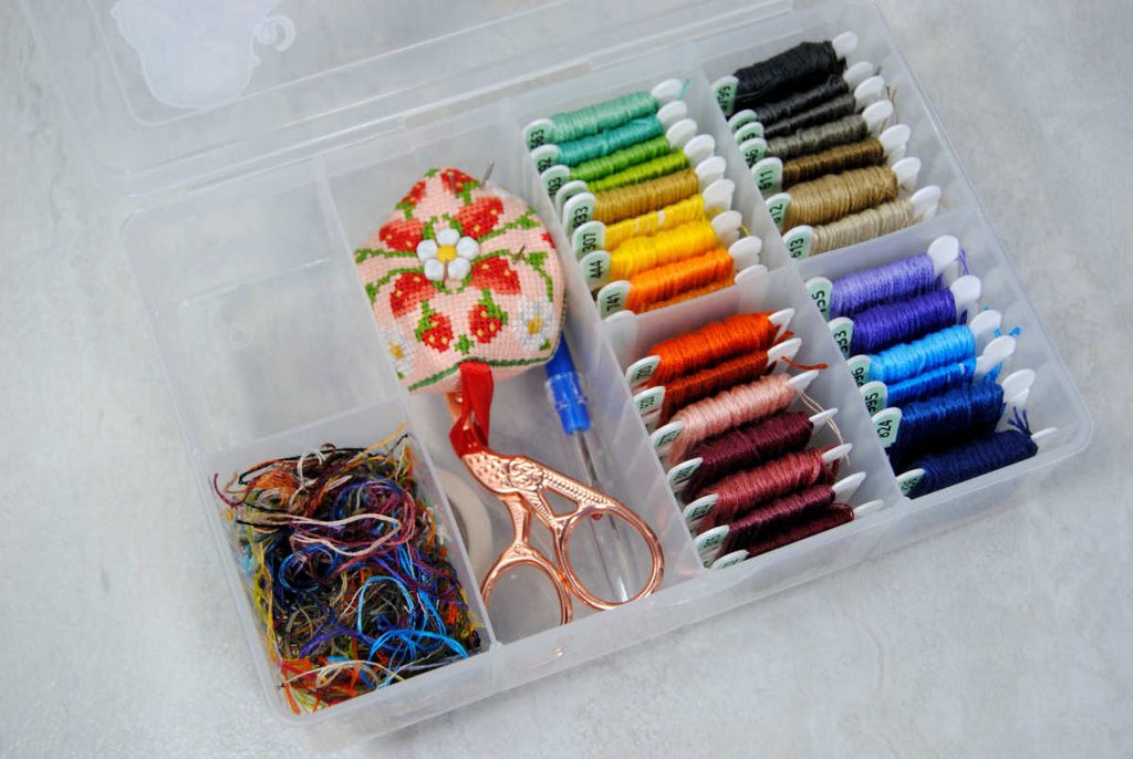 A small bobbin box full of embroidery thread on bobbins, a pincushion with needles, stork scissors, seam ripper, and a pile of ort threads.