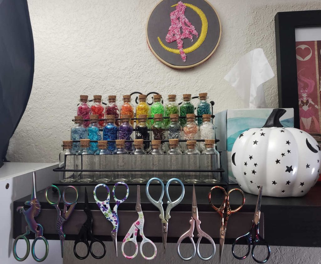 A display of thread-filled potion bottles behind a row of embroidery scissors stuck to a magnet strip on the edge of the shelf they're on.