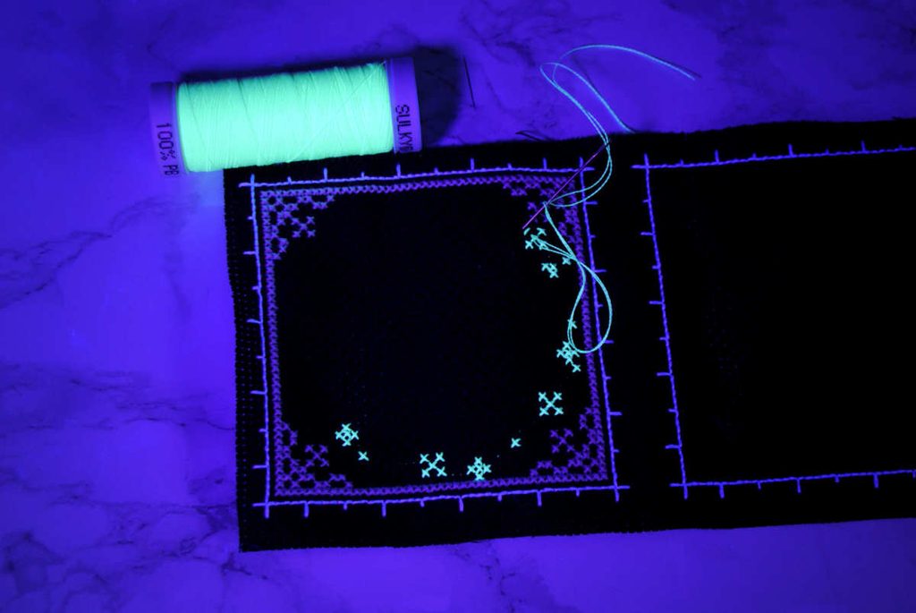 A partially stitched glow in the dark biscornu under blacklight