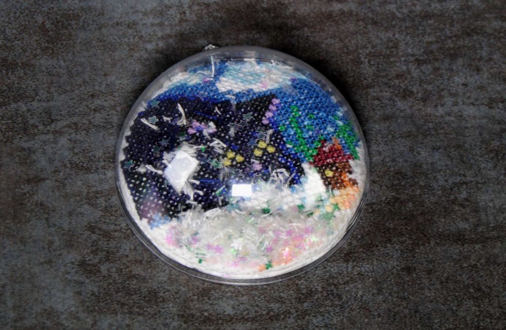 Close up view of the top half of a cross stitch snowglobe ornament full of glitter and thread snow.