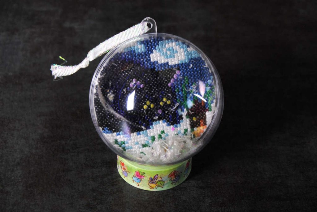 A cross stitched Yule cat in a snowglobe Christmas tree ornament on a washi tape stand.