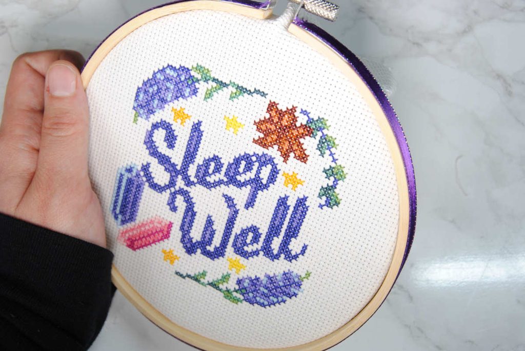A cross stitch project in an embroidery hoop embellished with ribbon along the outer edge.