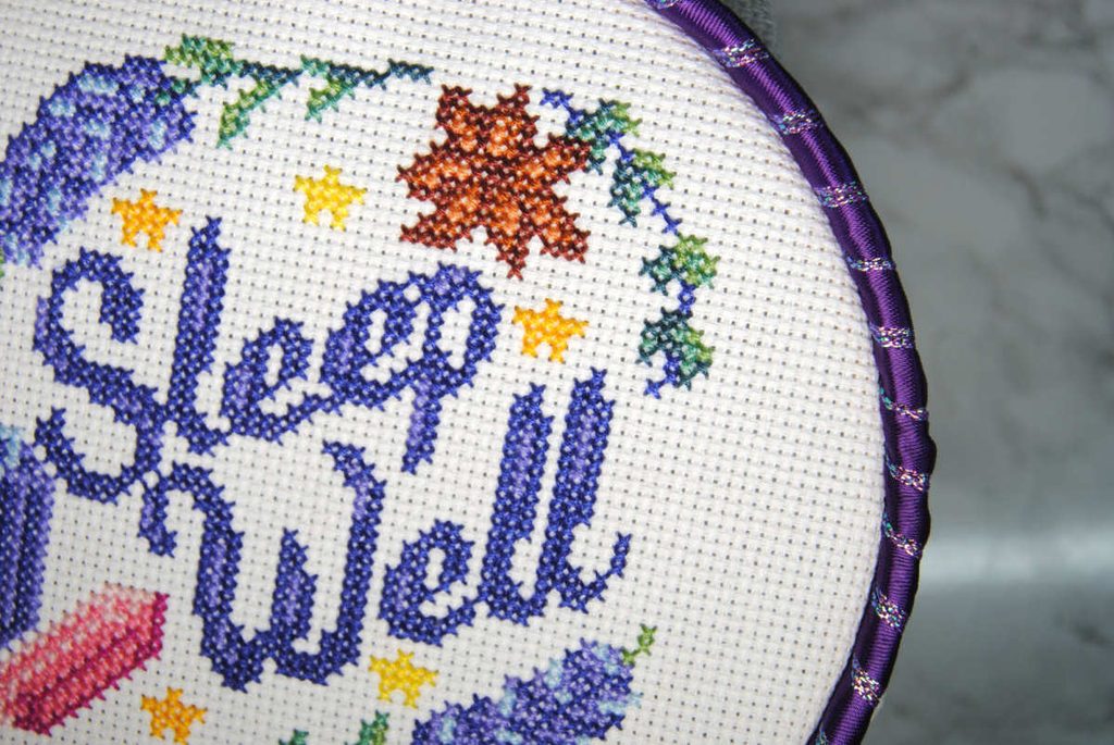 A cross stitch project in an embroidery hoop embellished with ribbon wrapped around the outer edge.