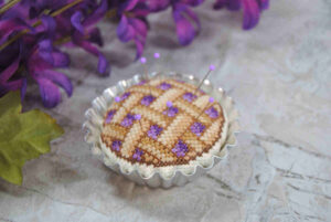 A cross stitch pin cushion resembling a blueberry pie in a tart tin.