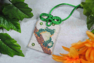 A cross stitch omamori charm featuring a frog, meant to ward off miscounts and frogging.